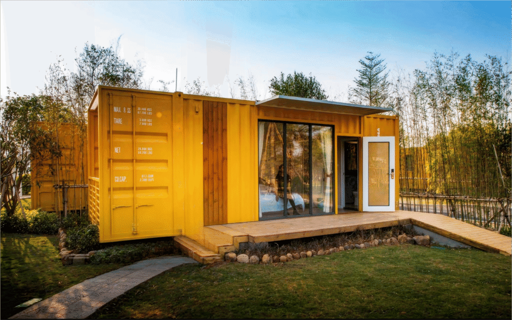 Light yellow shipping container tiny home with large glass sliding door, small front porch, surrounded by greenery and trees, showcasing modular prefabricated structure and eco-friendly construction.