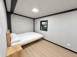 Sypialnia w rozkładanym domu kontenerowym 20ft z drewnianym łóżkiem, stolikiem nocnym i otwieranym oknem.