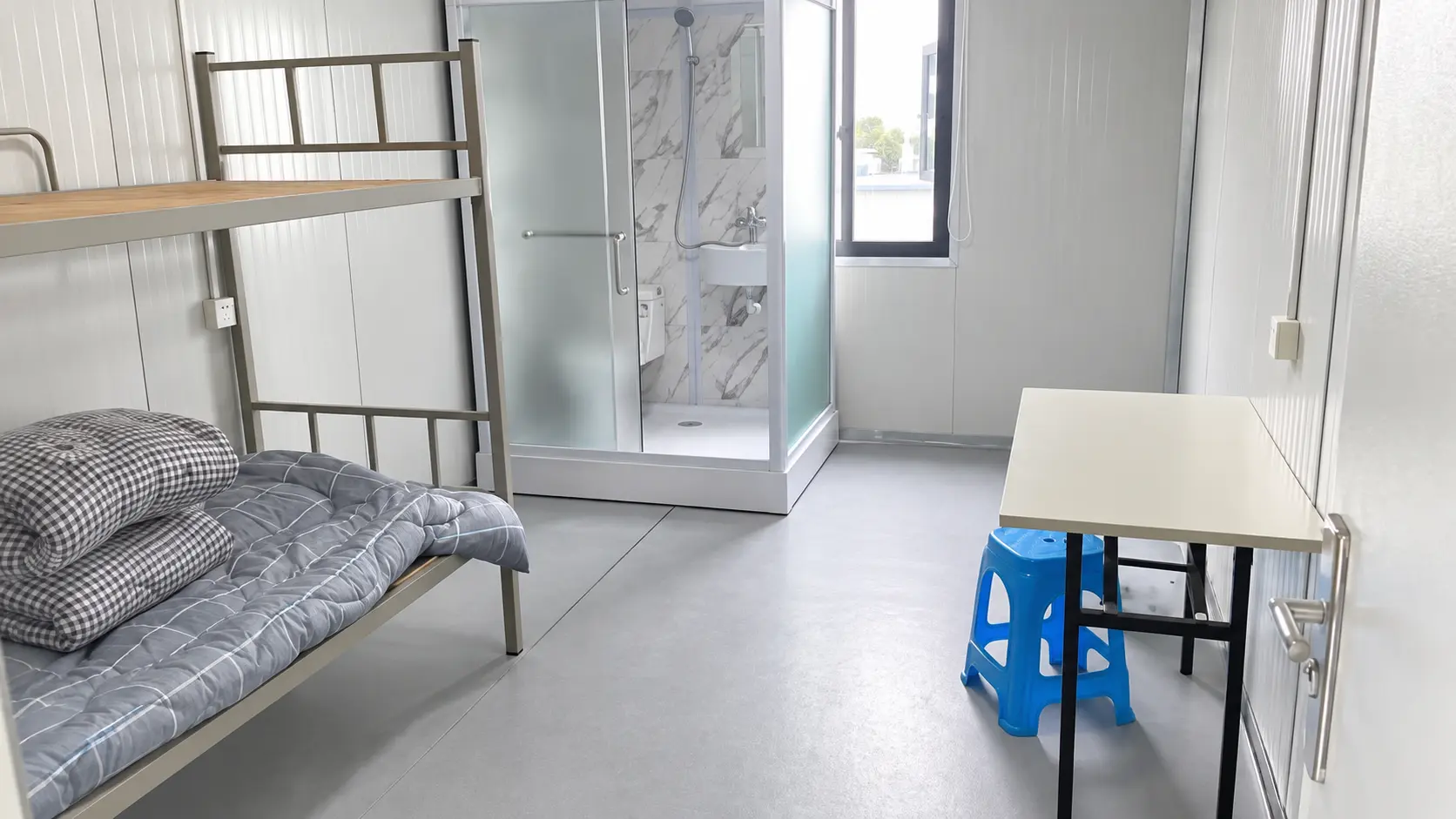 Flat-pack container dormitory interior with bunk bed, desk, chair and private shower area.