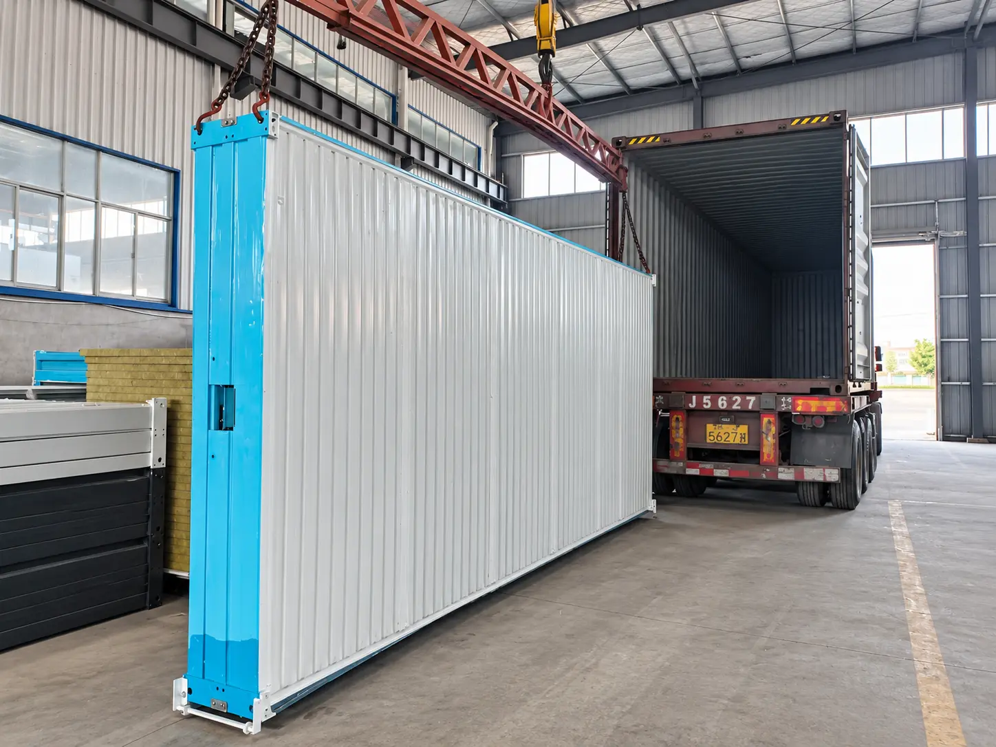 Factory crane handling a folded building assembly beside an open delivery truck